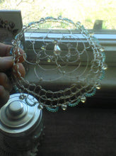 Load image into Gallery viewer, Silver and blue crystals on silver wire form a flower shaped kippah. The kippah is being held aganst a sunny window. There's a silver ldded jar in the foreground and a card in the background.