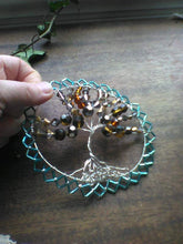 Load image into Gallery viewer, Tree of Life wall hanging in silver, amber, copper and blue being held on a brown wooden surface, with a vine in the background.