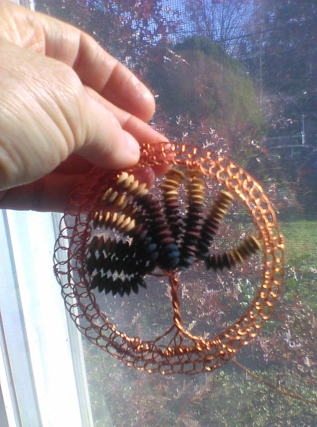 Bright copper wire supports wooden beads upoccled froma broken bracelet. Shown held againt a window, with autumn trees n the background.