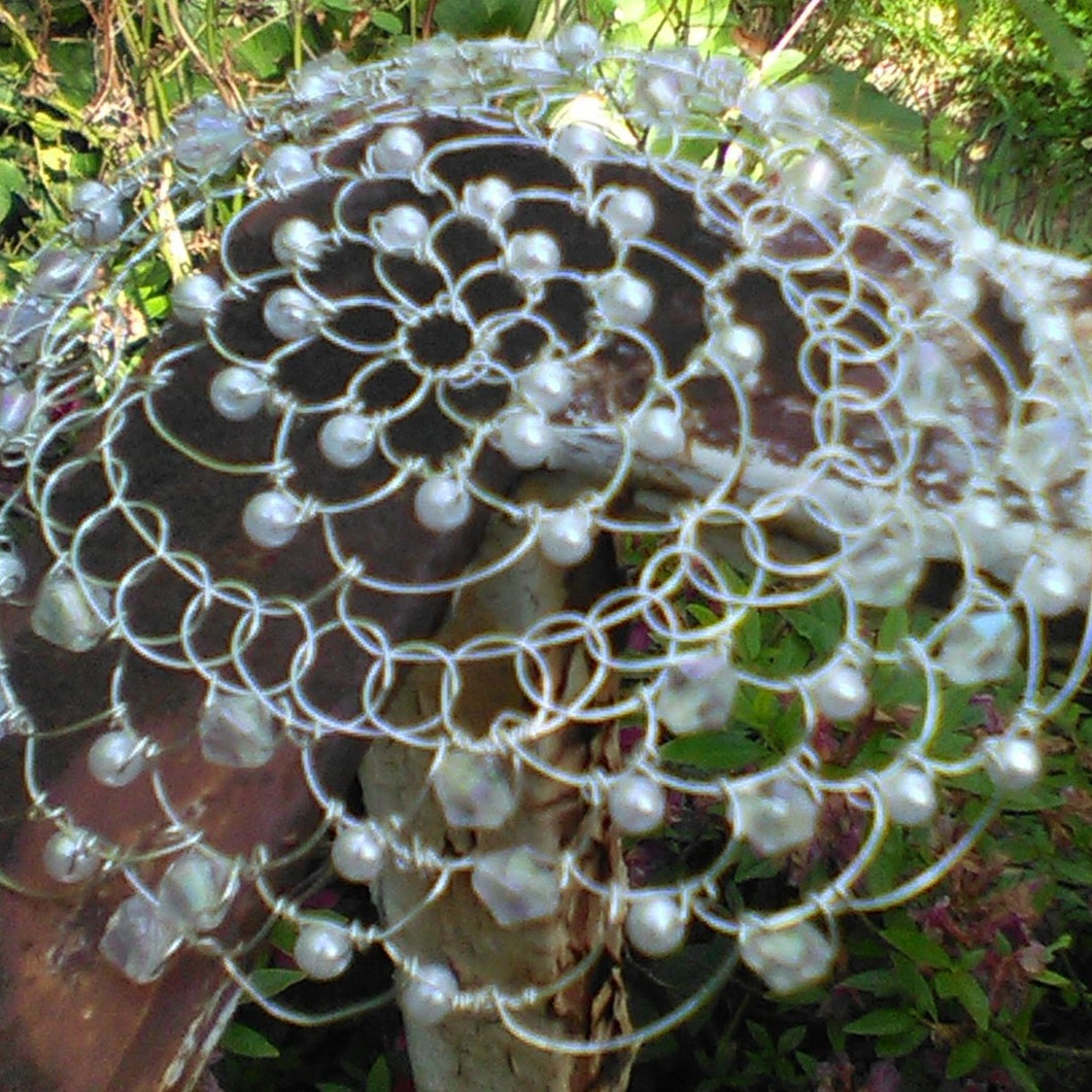 Crystals and pearls in abundance on silver wire kippah. The kippah is shown on a railing, wth green leaves in the background.